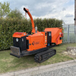 Orange Timberwolf wood chipper on tracks parked beside a hedge, with a tall metal fence and utility pole in the background.