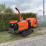 Orange Timberwolf wood chipper on tracks parked beside a hedge and metal fence under a cloudy sky in an outdoor industrial area