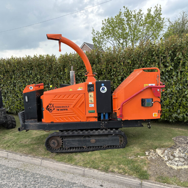 Orange Timberwolf TW 280TFTR wood chipper on tracks parked beside a tall hedge, with a cloudy sky above and a grassy verge nearby.