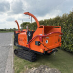 Orange tracked wood chipper on a grassy verge beside a hedge under a cloudy sky.