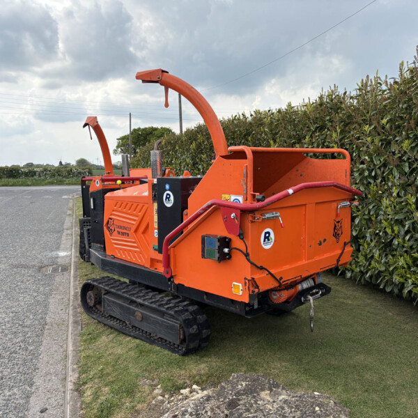 Orange tracked wood chipper on a grassy verge beside a hedge under a cloudy sky.