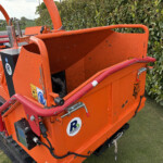 Orange wood chipper trailer parked by a hedge on a rural road, view from the side with guard rails and decals visible