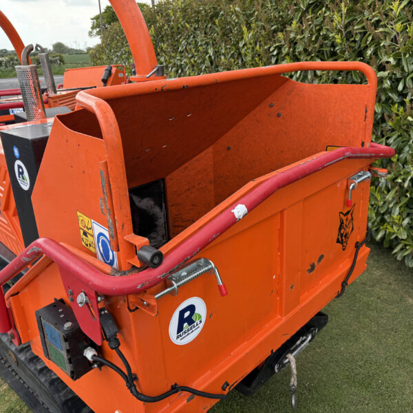 Orange wood chipper trailer parked by a hedge on a rural road, view from the side with guard rails and decals visible