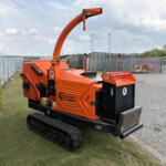 Orange Timberwolf wood chipper on a trailer in an outdoor yard with metal fencing and a parked car in the background.
