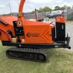 Orange Timberwolf TW 280FTFR wood chipper on crawler tracks in a yard, with a metal fence and parked vans in the background.