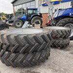 Stack of large, treaded tractor tires in a yard next to a blue tractor and workshop building.