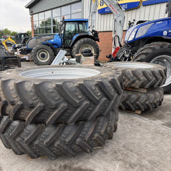 Stack of large, treaded tractor tires in a yard next to a blue tractor and workshop building.