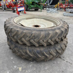 Two large tractor tires stacked on a paved surface with farm machinery in the background, including a green baler and hose reel.