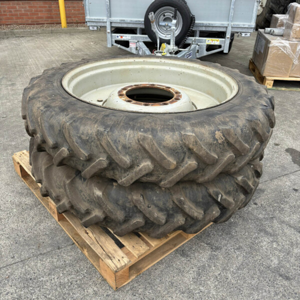 Large dirty tractor tire with a steel rim resting on a wooden pallet outdoors in a parking lot behind a trailer.