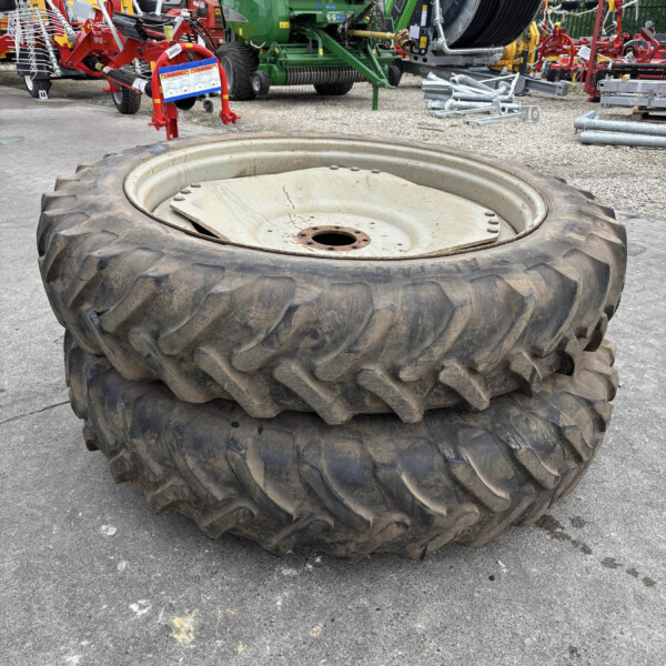 Two large tractor tires stacked on a paved surface with farm machinery in the background, including a green baler and hose reel.