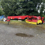 Red agricultural/industrial machine on a gravel lot with blue safety barriers and a dense tree line in the background, at a worksite edge.