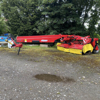 Red agricultural/industrial machine on a gravel lot with blue safety barriers and a dense tree line in the background, at a worksite edge.