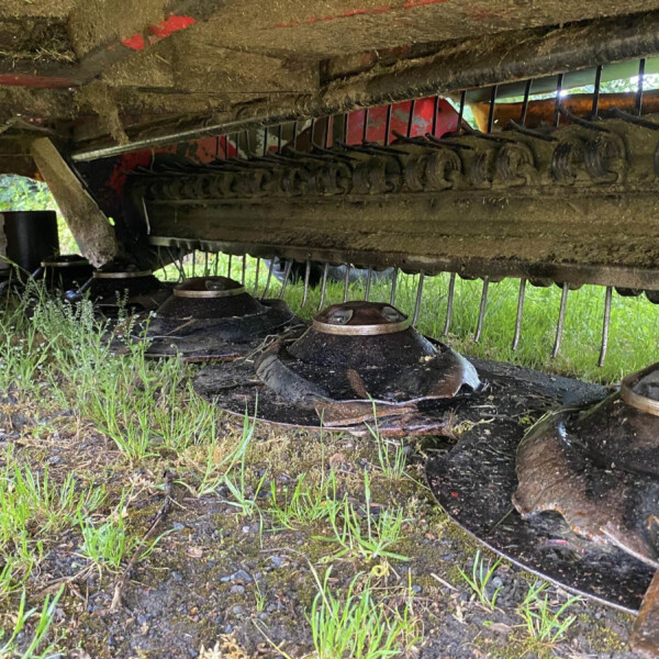 Underside of a muddy agricultural harvester header with a row of curved blades and guards, resting on grass and soil.
