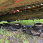 Underneath a dirty, rusted farming implement with rotating tines and curved blades, covered in soil and grass.