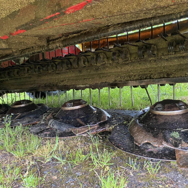 Underneath a dirty, rusted farming implement with rotating tines and curved blades, covered in soil and grass.