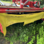 Close-up of a red farm mower attachment with a yellow cutting guard, resting in tall weeds.