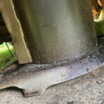 Close-up of a large, dark metal bell with a rusty strap, covered in dirt, outdoors among leaves and soil.
