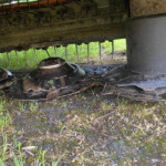 Underside of a dirty farming implement with row of metal tines and grime, seen from below.