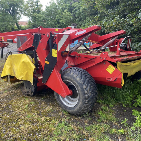 Red agricultural machine with large tires and yellow protective covers parked on a gravel lot beside trees and bushes, ready for use