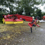 Red agricultural machine with a long arm resting in a gravel lot, trees in the background, likely farming equipment awaiting use.