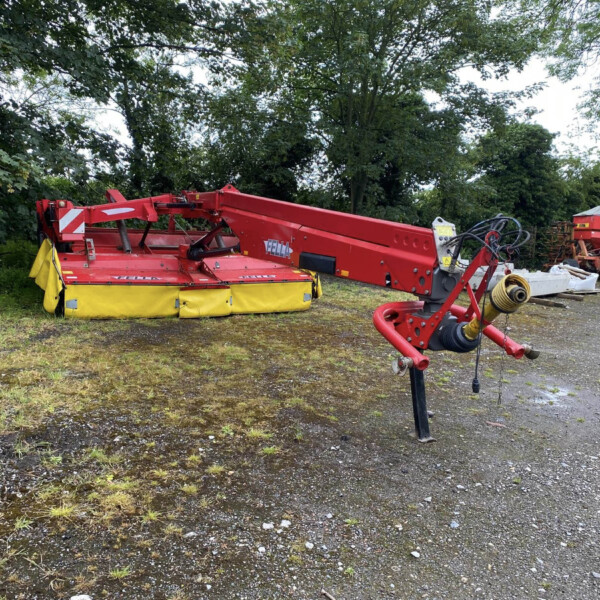 Red agricultural machine with a long arm resting in a gravel lot, trees in the background, likely farming equipment awaiting use.