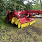 Red agricultural mower/baler machine parked in a grassy, leafy area with a yellow protective cover.