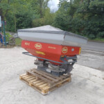 Red and gray boat trailer on a pallet in a parking lot near parked cars and green trees, with a gray cover over the boat hull.
