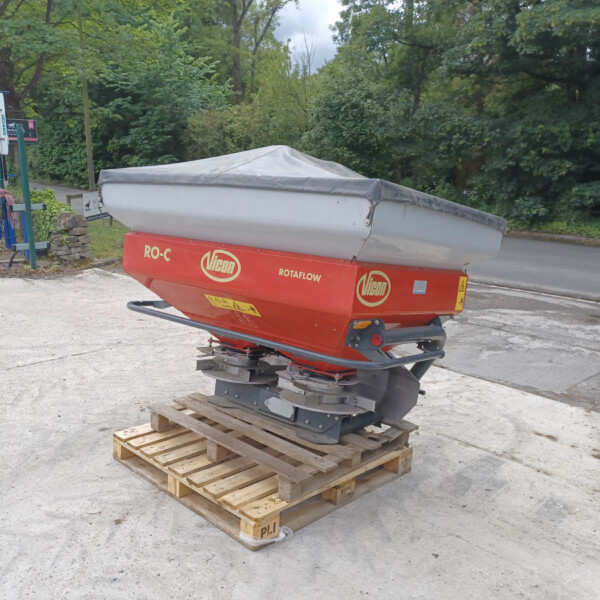 Red and gray boat trailer on a pallet in a parking lot near parked cars and green trees, with a gray cover over the boat hull.