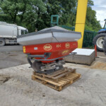 Red Vicon RO-C Rotaflow spreader on a wooden pallet in an outdoor yard, with a white truck and a blue tractor in the background.