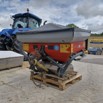 Blue tractor cab with attached red farming implement resting on wooden pallets in a parking lot near a farm supply store.