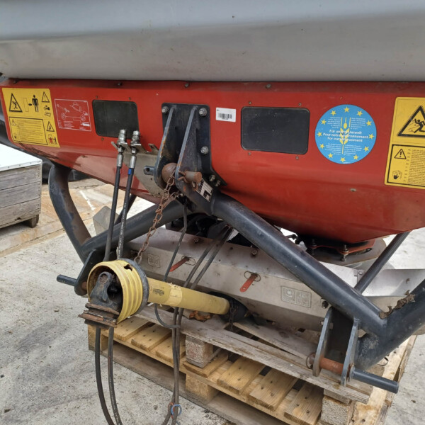 Close-up of a red agricultural machine base with yellow warning labels, black access panels, and connected hoses/chains on a wooden pallet.