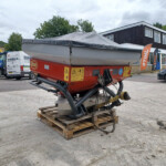 Red boat on a trailer sits on wooden pallets in an outdoor industrial yard, with vans and a dark building in the background.