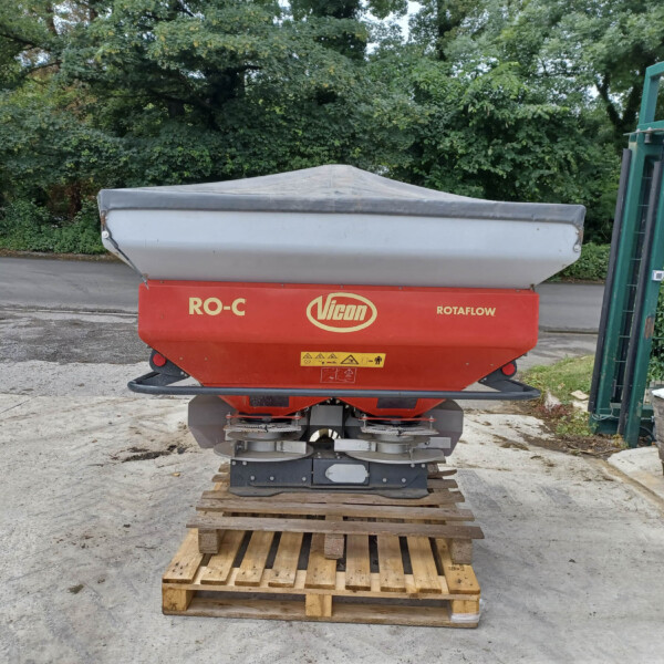 Red Vicon ROTAFLOW agricultural spreader on wooden pallets in an outdoor yard, with a gray tarp cover and green fence in the background.