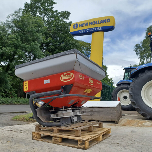 Tractor attached to a red agricultural machine on a wooden pallet outdoors under a blue and yellow New Holland Agriculture sign.