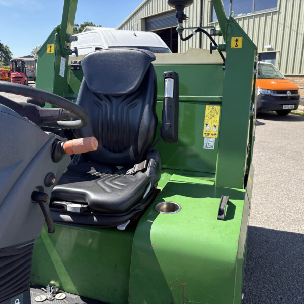 Close-up of a green agricultural machine cab with black seat, joystick, and control panels outdoors in a yard with vans and equipment in the background.