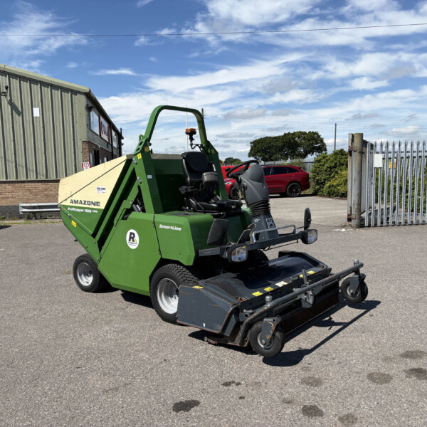 Green agricultural implement (Amazone) in a parking lot, with a red car, metal fence, and warehouse in the background under a blue sky.