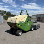 Green Amazone Profihopper 1500 farming trailer in a paved yard with a metal fence and blue sky behind it, partially loaded with a beige hopper.