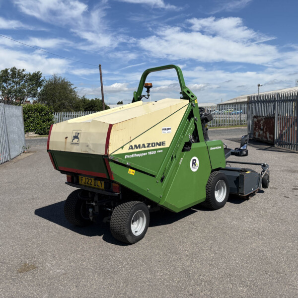Green Amazone Profihopper 1500 farming trailer in a paved yard with a metal fence and blue sky behind it, partially loaded with a beige hopper.