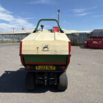 Rear view of a beige-and-green dump trailer with an orange warning beacon, parked in an industrial yard next to a red skip bin and metal fencing.