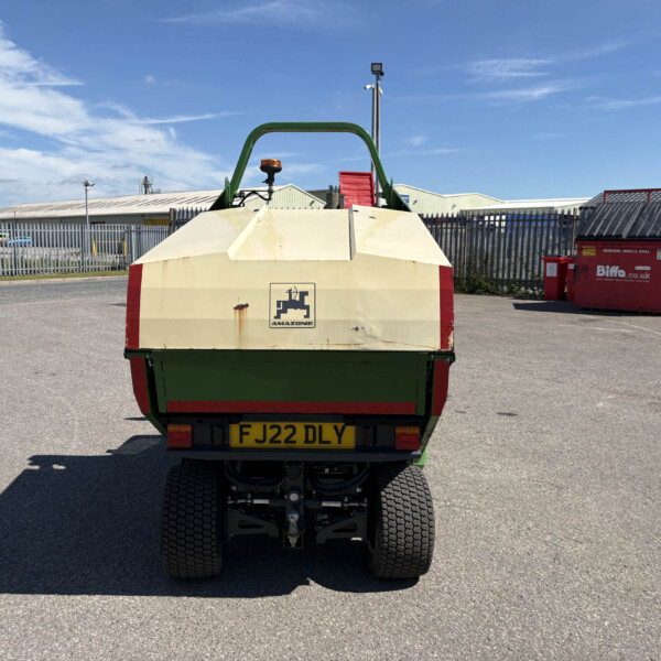 Rear view of a beige-and-green dump trailer with an orange warning beacon, parked in an industrial yard next to a red skip bin and metal fencing.