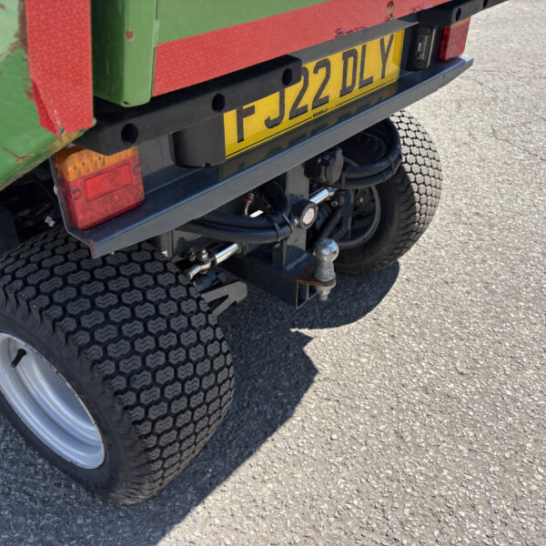 Rear view of a green and red truck trailer showing a yellow license plate with black characters, plus the dual-tire axle and towing hardware visible at the back of the vehicle.