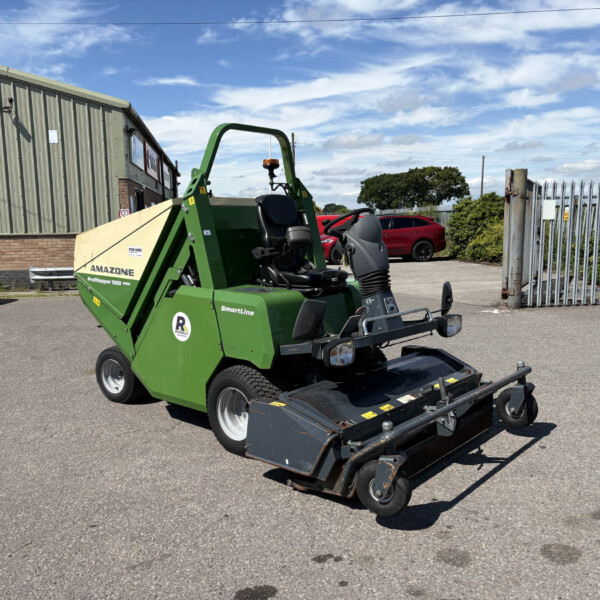 Green industrial machine with a front attachment parked in a paved lot beside a corrugated metal building under a blue sky.