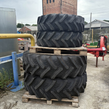 Stack of large tractor tires stacked on wooden pallets in an outdoor industrial yard with a brick tower in the background when viewed from the side.