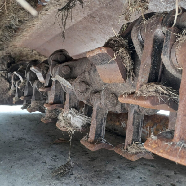 Close-up of a rusted metal undercarriage with wheels or linked gears, covered in dust and dried plant fibers, seen from the ground level