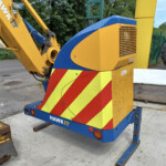 Back side of a yellow construction machine with red-and-yellow diagonal hazard stripes on a blue trailer, parked on a street with greenery in the background.