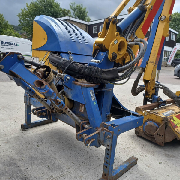 Close-up of a blue and yellow hydraulic construction machine with exposed hoses and joints, outdoors in a yard with vans in the background.