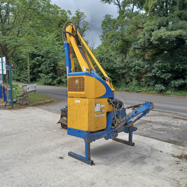 Yellow and blue construction machine sits on a paved area beside a road, with green trees in the background.