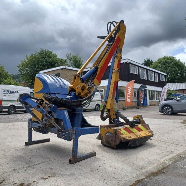 Construction excavator with yellow and blue arms in a parking lot under a cloudy sky, with service vans in the background.