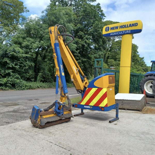 Construction excavator with yellow and blue arms mounted on a wheeled trailer beside a New Holland Agriculture sign, outdoors under trees.