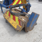 Close-up of a weathered construction bucket with yellow and red chevron safety markings and blue metal framework, showing worn paint and hydraulic hoses.